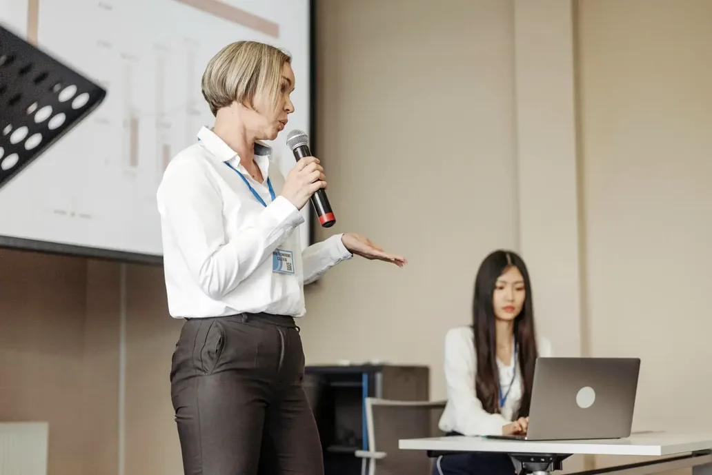 A woman with a microphone presents in front of a screen while a colleague works on a laptop beside her