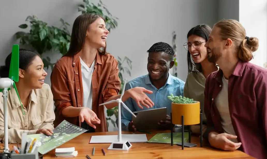 A laughing group of young people around a table with a small wind turbine model, a tablet and a potted plant