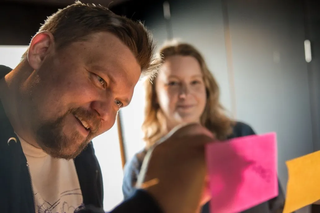 Two MinnaLearn team members smiling as they work with pink and yellow sticky notes on a glass wall