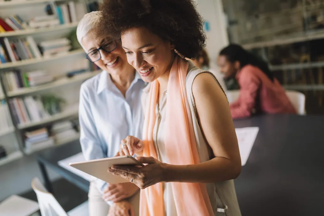 Two smiling women look at a tablet together in a library-like office
