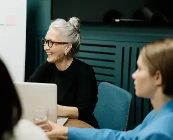 A smiling woman with gray hair and glasses talking with colleagues at a table